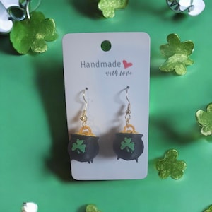 Può includere: Orecchini fatti a mano con ciondoli a forma di pentola neri, ciascuno con un trifoglio verde. Gli orecchini pendono da ganci color oro e sono esposti su una carta bianca con la scritta "Handmade with love". Lo sfondo è verde con decorazioni a trifoglio.