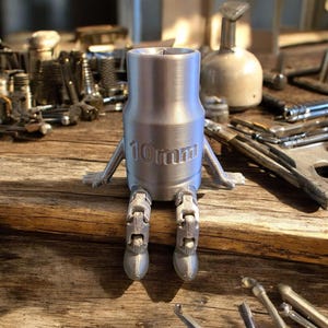 Puede incluir: Una llave de vaso plateada de 10 mm con brazos y piernas adjuntos, lo que le da una apariencia humanoide. La llave está sobre una superficie de madera, con varias herramientas en el fondo.