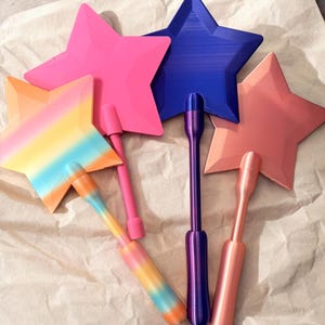 May include: A collection of colorful star-shaped wands in shades of pink, blue, peach, and rainbow. Each wand features a star top and a handle. The wands are made of plastic and are arranged on a light brown surface.