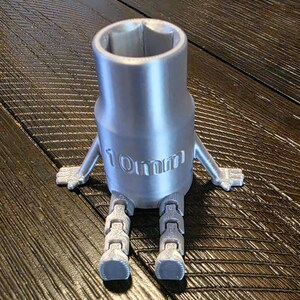 Puede incluir: Una llave de vaso plateada de 10 mm con piernas y brazos articulados, lo que le da una apariencia de dibujos animados. El texto "10mm" está impreso en el lateral. La llave está sobre una superficie de madera oscura.
