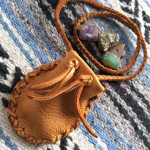 May include: A small, tan leather pouch with braided edges and a drawstring closure. A braided leather cord is attached. The pouch is next to several small, colorful gemstones, including amethyst and turquoise.