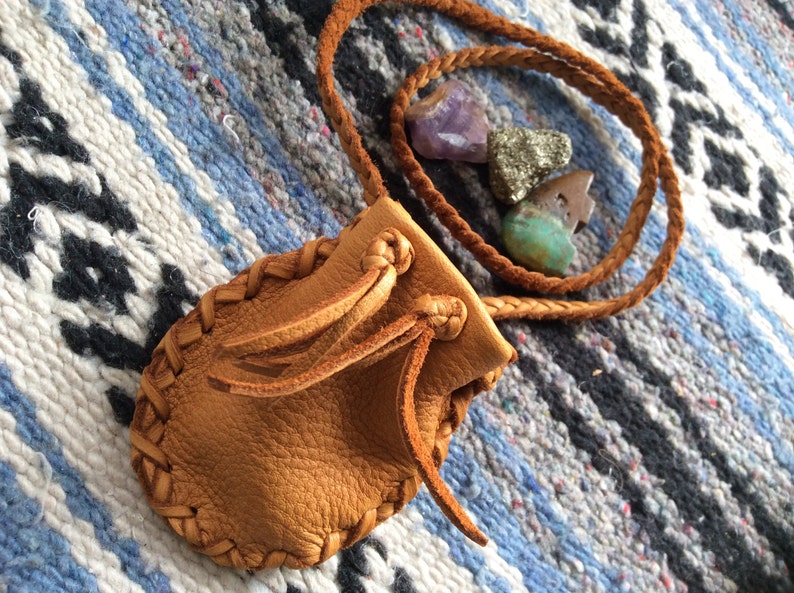 May include: A small, tan leather pouch with braided edges and a drawstring closure. A braided leather cord is attached. The pouch is next to several small, colorful gemstones, including amethyst and turquoise.