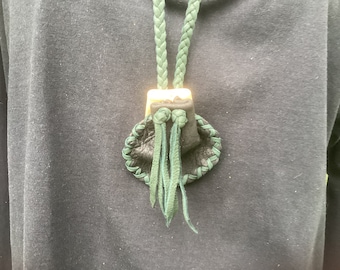 May include: A small black leather pouch with green leather trim and tassels hangs from a green braided cord. The pouch is closed with a gold metal clasp.