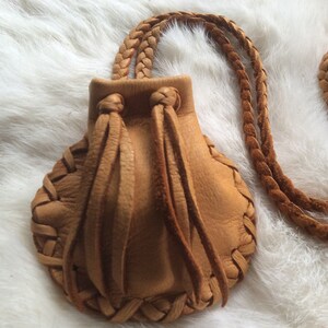 May include: A tan leather pouch necklace with a braided strap. The pouch has a scalloped edge and is gathered at the top with leather ties. The necklace is made of braided leather.