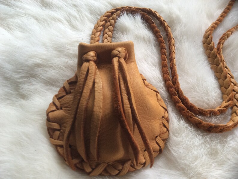 May include: A tan leather pouch necklace with a braided strap. The pouch has a scalloped edge and is gathered at the top with leather ties. The necklace is made of braided leather.