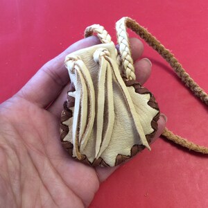 May include: A small, handmade leather pouch with a braided strap. The pouch is cream-colored with a brown, scalloped edge and decorative fringe. The strap is a twisted, light brown cord.