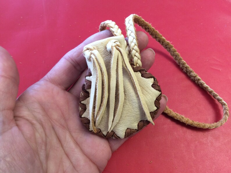 May include: A small, handmade leather pouch with a braided strap. The pouch is cream-colored with a brown, scalloped edge and decorative fringe. The strap is a twisted, light brown cord.