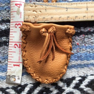 May include: A small, tan leather pouch with a drawstring closure and braided edges. The pouch is approximately 3 inches (7.6 cm) tall and 2 inches (5 cm) wide. A measuring tape and ruler are next to the pouch.