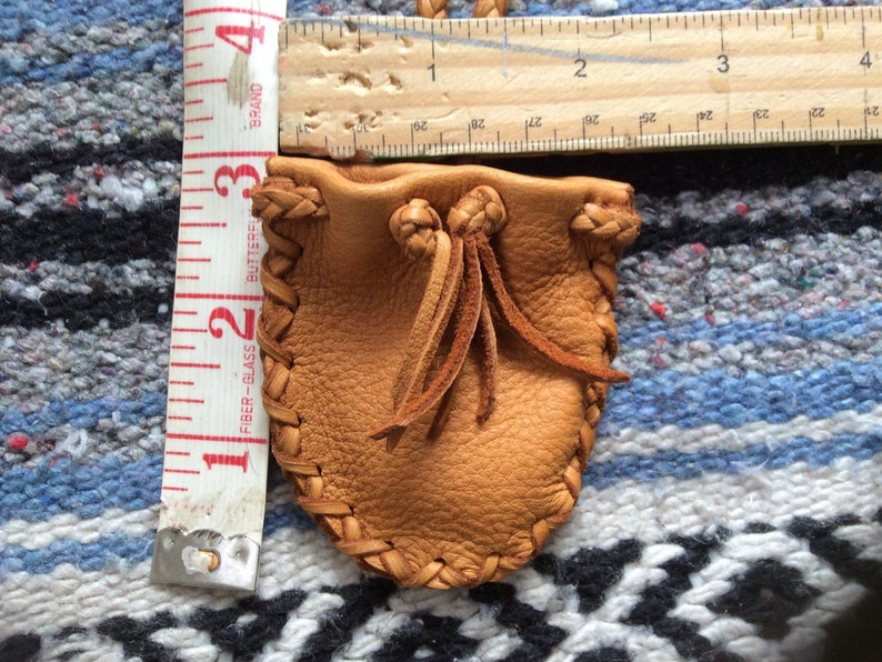 May include: A small, tan leather pouch with a drawstring closure and braided edges. The pouch is approximately 3 inches (7.6 cm) tall and 2 inches (5 cm) wide. A measuring tape and ruler are next to the pouch.