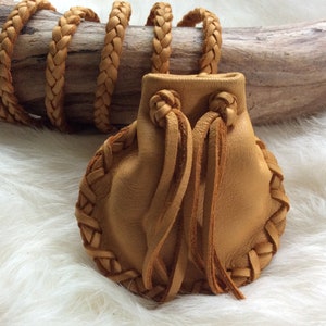 K&ouml;nnte beinhalten: Eine hellbraune Ledertasche mit geflochtenem Rand und Kordelzug. Die Tasche wird von einer langen, geflochtenen Lederschnur gehalten. Der Hintergrund zeigt ein St&uuml;ck Treibholz und eine wei&szlig;e, flauschige Oberfl&auml;che.