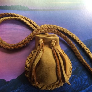May include: A tan leather pouch necklace with a braided cord. The small pouch has a rounded shape with decorative stitching and fringe. The background is a colorful landscape with shades of blue, purple, and green.