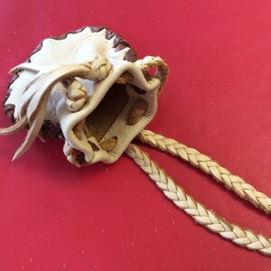 May include: A handmade leather pouch with a drawstring closure and braided cord. The pouch is cream-colored with a dark brown trim and decorative knots. The background is a solid red.