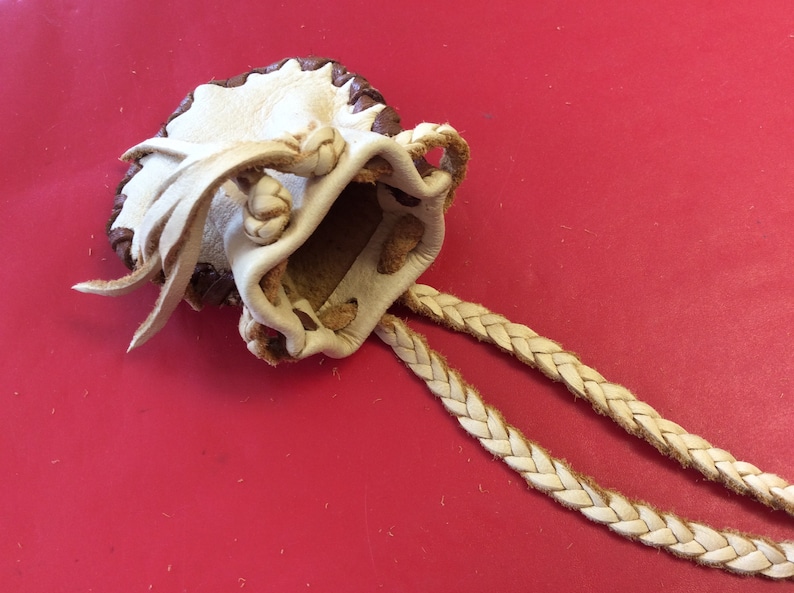 May include: A handmade leather pouch with a drawstring closure and braided cord. The pouch is cream-colored with a dark brown trim and decorative knots. The background is a solid red.