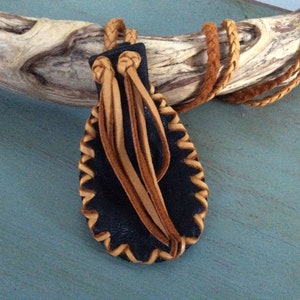May include: A black leather pouch with a tan leather braided cord and fringe. The pouch is oval shaped and has a decorative stitch around the edge.