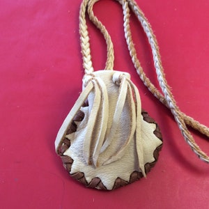 May include: A small, cream-colored leather pouch with a braided strap. The pouch has a scalloped edge with brown stitching and decorative fringe. The background is a solid red.