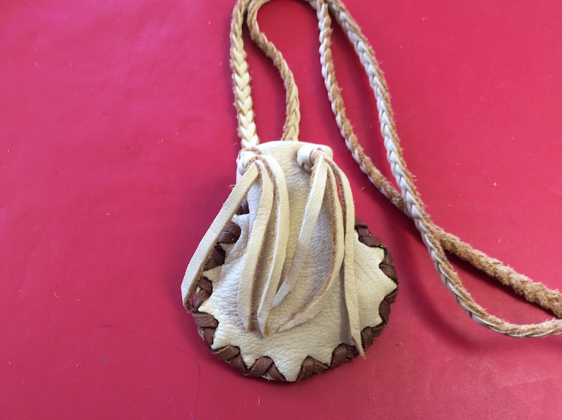 May include: A small, cream-colored leather pouch with a braided strap. The pouch has a scalloped edge with brown stitching and decorative fringe. The background is a solid red.