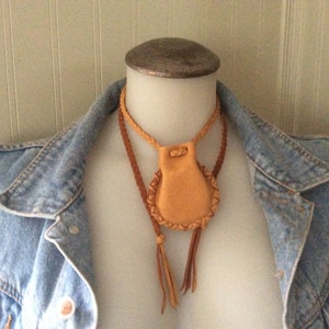 May include: A tan leather pouch necklace with a braided leather cord and tassels. The pouch has a stitched border and a drawstring closure. The necklace is displayed on a mannequin wearing a denim jacket.