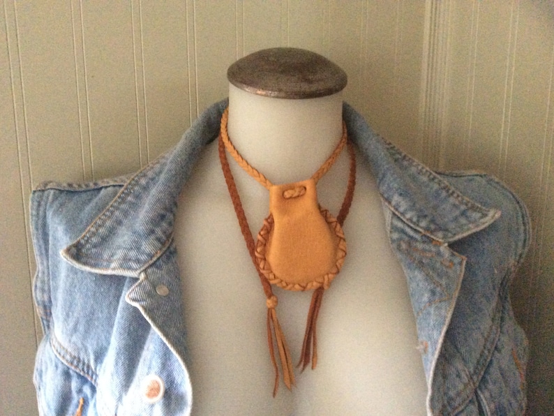 May include: A tan leather pouch necklace with a braided leather cord and tassels. The pouch has a stitched border and a drawstring closure. The necklace is displayed on a mannequin wearing a denim jacket.