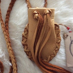 May include: A handcrafted leather pouch with a braided strap. The pouch is tan with braided edges and decorative tassels. The strap is also braided. A tag with the word "Leather" is attached.