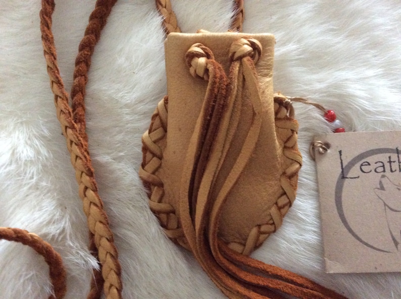 May include: A handcrafted leather pouch with a braided strap. The pouch is tan with braided edges and decorative tassels. The strap is also braided. A tag with the word "Leather" is attached.