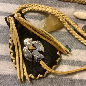 Könnte beinhalten: Eine schwarze Ledertasche mit silbernem Emblem, beigefarbenen Fransen und einem geflochtenen beigefarbenen Riemen. Die Tasche ist mit einem silbernen Emblem verziert. Zusätzliche dekorative Elemente sind eine fischförmige Schnitzerei und verschiedene Edelsteine.