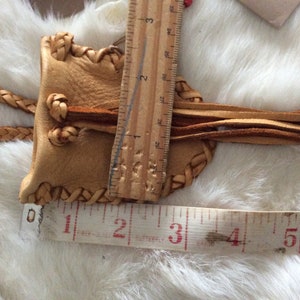 May include: A tan leather bolo tie slide with braided edges and a braided cord. The slide is approximately 2 inches long. Tassels hang from the bottom of the slide. The bolo tie is on a white furry surface.