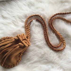 May include: A tan leather pouch with a braided strap and drawstring closure. The pouch has a fringed edge and is set against a white, fluffy background. The leather appears soft and worn.