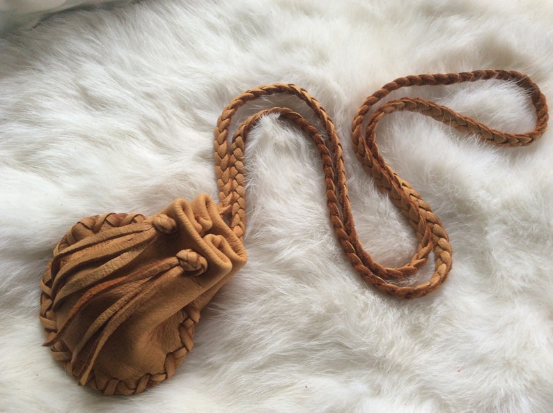 May include: A tan leather pouch with a braided strap and drawstring closure. The pouch has a fringed edge and is set against a white, fluffy background. The leather appears soft and worn.