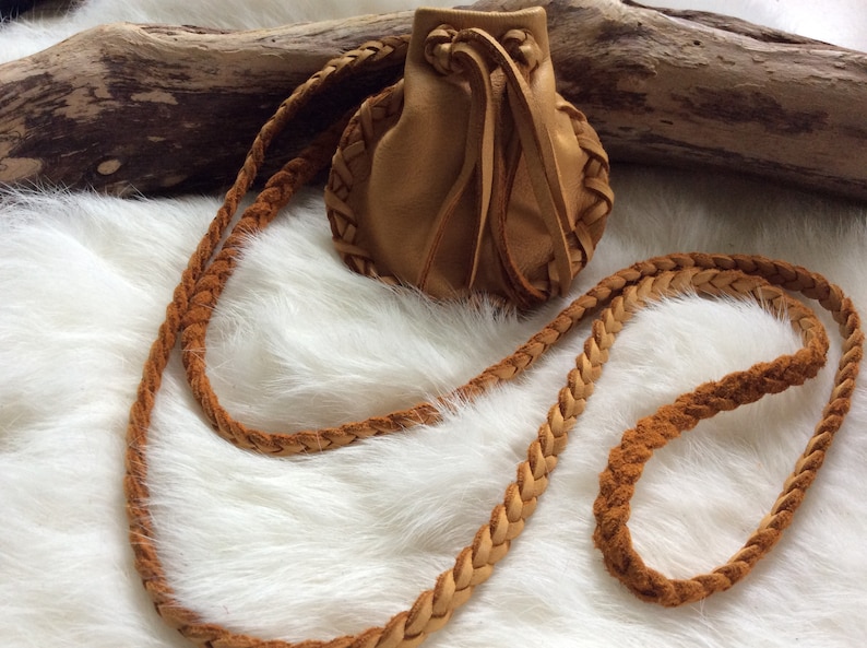 K&ouml;nnte beinhalten: Eine kleine, hellbraune Ledertasche mit geflochtenem Riemen. Die Tasche hat einen Kordelzugverschluss und ist mit geflochtenen Lederakzenten verziert. Der Riemen ist ebenfalls geflochten. Der Beutel liegt auf einem wei&szlig;en, flauschigen Hintergrund.