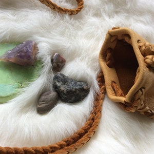 K&ouml;nnte beinhalten: Eine hellbraune Ledertasche mit geflochtenen Verzierungen und einem geflochtenen Riemen, zusammen mit verschiedenen Steinen und einem Amethystst&uuml;ck auf einer wei&szlig;en, fellartigen Oberfl&auml;che. Die Tasche ist offen.