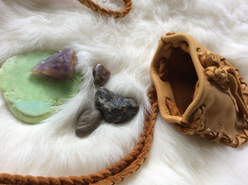 K&ouml;nnte beinhalten: Eine hellbraune Ledertasche mit geflochtenen Verzierungen und einem geflochtenen Riemen, zusammen mit verschiedenen Steinen und einem Amethystst&uuml;ck auf einer wei&szlig;en, fellartigen Oberfl&auml;che. Die Tasche ist offen.
