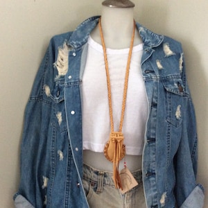 May include: A light brown leather necklace with a small pouch pendant and a braided cord. The necklace is displayed on a white top and a distressed denim jacket. The jacket has ripped details.
