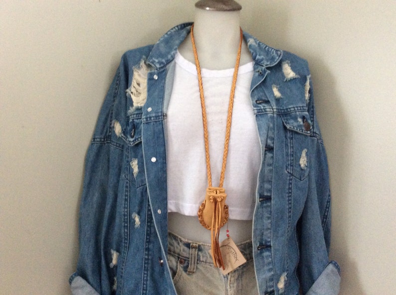 May include: A light brown leather necklace with a small pouch pendant and a braided cord. The necklace is displayed on a white top and a distressed denim jacket. The jacket has ripped details.