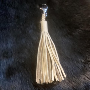 May include: A cream-colored leather tassel with a silver-tone clasp. The tassel is made of multiple strips of leather and hangs against a dark, textured background. This accessory can be used as a keychain or bag charm.