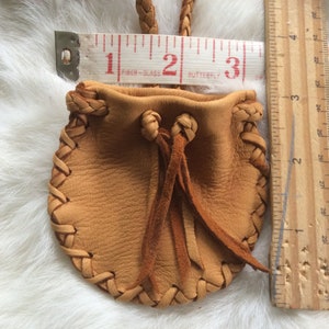 May include: A small, tan leather pouch with a braided drawstring and a braided strap. The pouch is approximately 7.6 cm wide and 7.6 cm tall. The edges are stitched with a decorative braid.