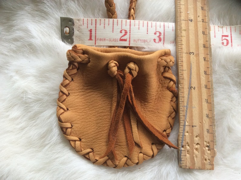 May include: A small, tan leather pouch with a braided drawstring and a braided strap. The pouch is approximately 7.6 cm wide and 7.6 cm tall. The edges are stitched with a decorative braid.