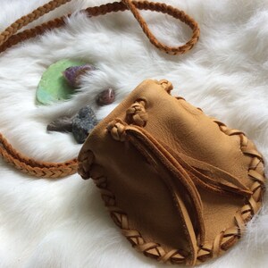 K&ouml;nnte beinhalten: Ein hellbrauner Lederbeutel mit geflochtenem Riemen und dekorativen N&auml;hten. Die kleine Tasche hat einen Kordelzugverschluss und wird auf einer wei&szlig;en, flauschigen Oberfl&auml;che pr&auml;sentiert. Kleine Steine und ein gr&uuml;ner Stein liegen neben dem Beutel.