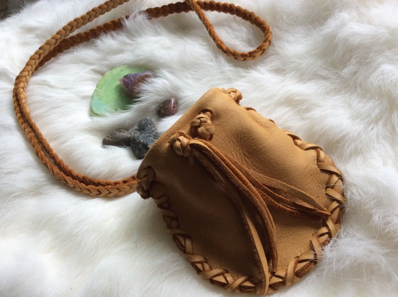 K&ouml;nnte beinhalten: Ein hellbrauner Lederbeutel mit geflochtenem Riemen und dekorativen N&auml;hten. Die kleine Tasche hat einen Kordelzugverschluss und wird auf einer wei&szlig;en, flauschigen Oberfl&auml;che pr&auml;sentiert. Kleine Steine und ein gr&uuml;ner Stein liegen neben dem Beutel.