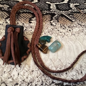 Op de afbeelding: Een bruine leren beurs met een gevlochten koord en kwastjes. De beurs ligt op een oppervlak met een slangenhuidpatroon met drie edelstenen: een groene tijgeroog, een lichtgroene steen en een donkergroene steen.