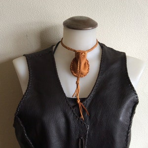 May include: A brown leather necklace with a small pouch pendant and a braided cord. The pendant is round with a tassel. The necklace is displayed on a mannequin wearing a dark brown leather vest.