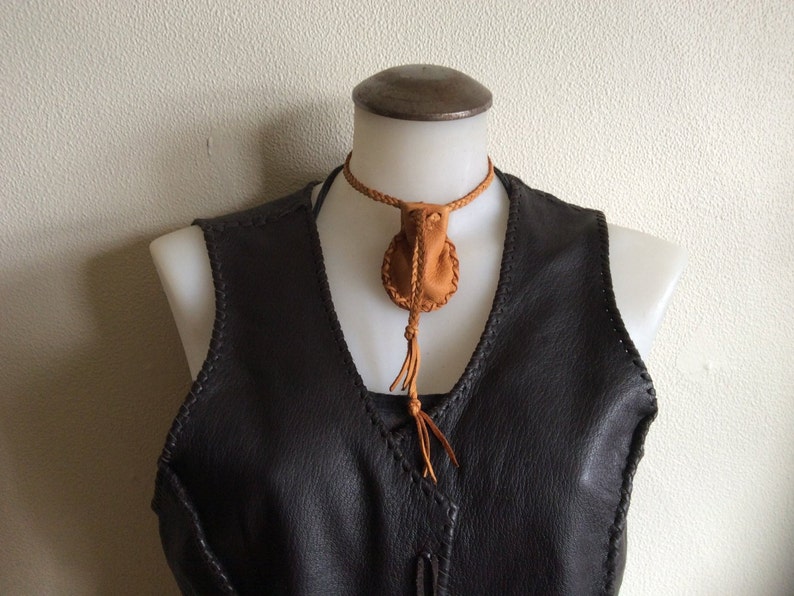 May include: A brown leather necklace with a small pouch pendant and a braided cord. The pendant is round with a tassel. The necklace is displayed on a mannequin wearing a dark brown leather vest.