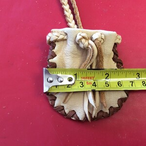 May include: A small, handmade leather pouch with a braided cord. The pouch is cream-colored with brown stitching and trim. It measures approximately 3 inches in length.