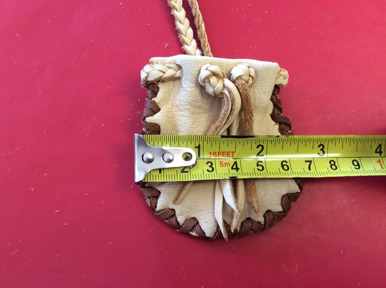 May include: A small, handmade leather pouch with a braided cord. The pouch is cream-colored with brown stitching and trim. It measures approximately 3 inches in length.