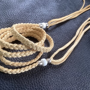 Op de afbeelding: Gevlochten leren wikkelarmband in een natuurlijke bruintint. De armband is opgerold en heeft een reeks gevlochten leren strengen. De uiteinden van de armband hebben lange, dunne leren bandjes en kleine zilveren kralen.