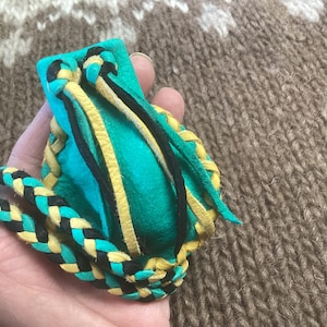 Peut inclure: Une pochette en cuir turquoise avec des bordures tressées jaunes et noires. La pochette est fermée par un cordon tressé.