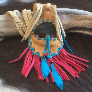 May include: A small, tan leather pouch with a blue and white trim. The pouch has a black leather flap and is decorated with red and blue fringe. The pouch is hanging from a braided white cord.