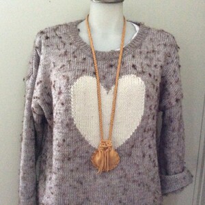K&ouml;nnte beinhalten: Eine hellbraune Lederkette mit einem kleinen Beutelanh&auml;nger h&auml;ngt &uuml;ber einem grauen Pullover mit einem gro&szlig;en wei&szlig;en Herzdesign. Der Pullover hat ein gesprenkeltes Muster und hochgekrempelte &Auml;rmel, was einen l&auml;ssigen, handgemachten Look erzeugt.