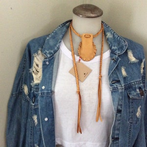 May include: A tan leather necklace with a braided cord and a small pouch pendant. The necklace is displayed on a mannequin wearing a white t-shirt and a distressed denim jacket. A small tag hangs from the necklace.