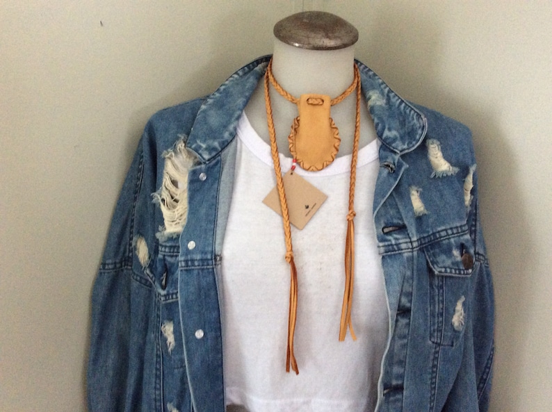 May include: A tan leather necklace with a braided cord and a small pouch pendant. The necklace is displayed on a mannequin wearing a white t-shirt and a distressed denim jacket. A small tag hangs from the necklace.