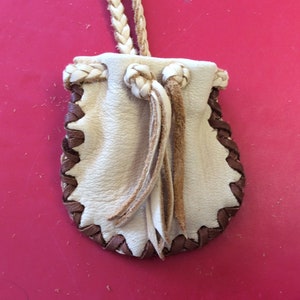 May include: A small, cream-colored leather pouch with a braided drawstring and a brown, stitched border. The pouch has a braided strap for carrying. The design is simple and rustic, suitable for small items.